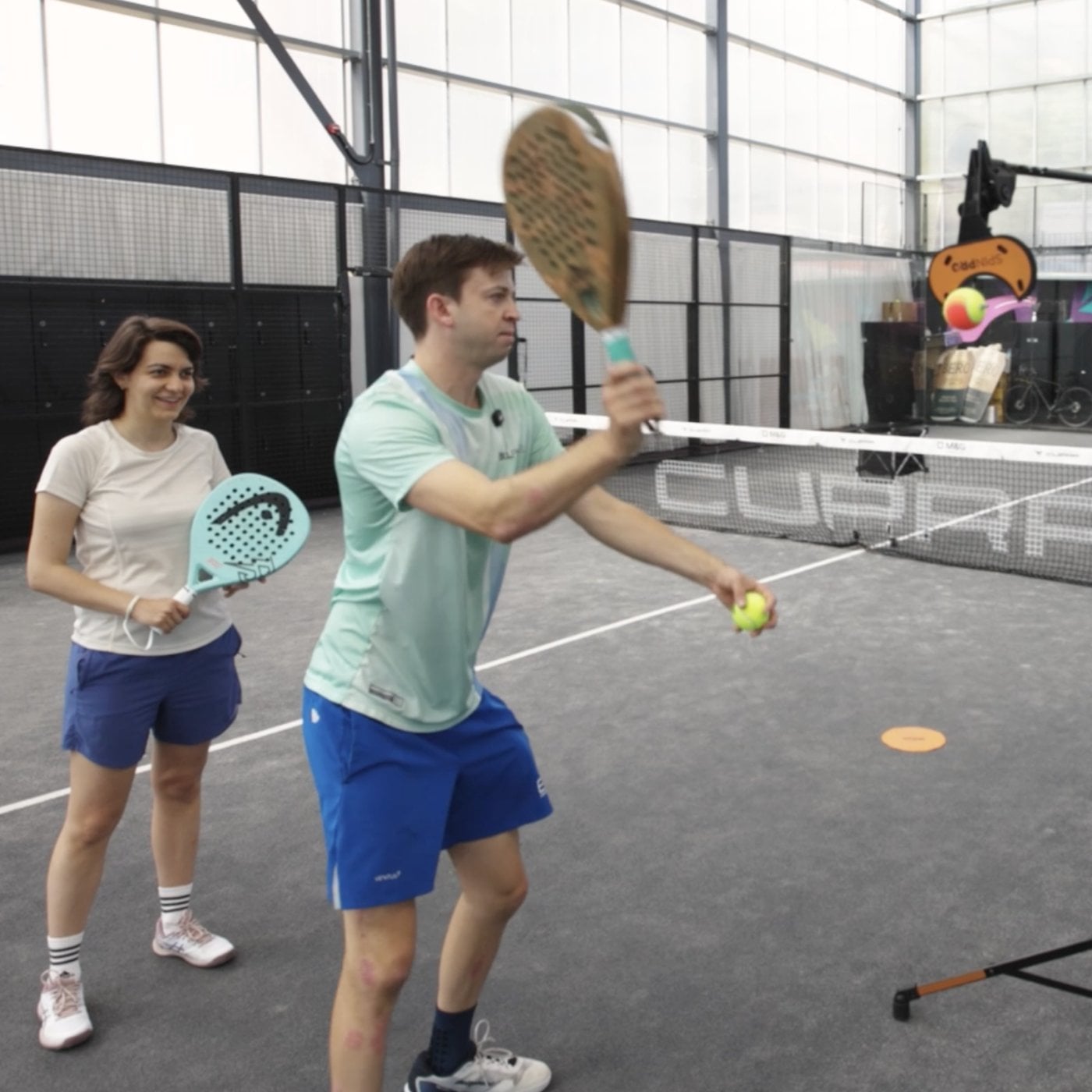 A coach demonstrating a shot to a player on a padel court, with the SpinPro set up behind them