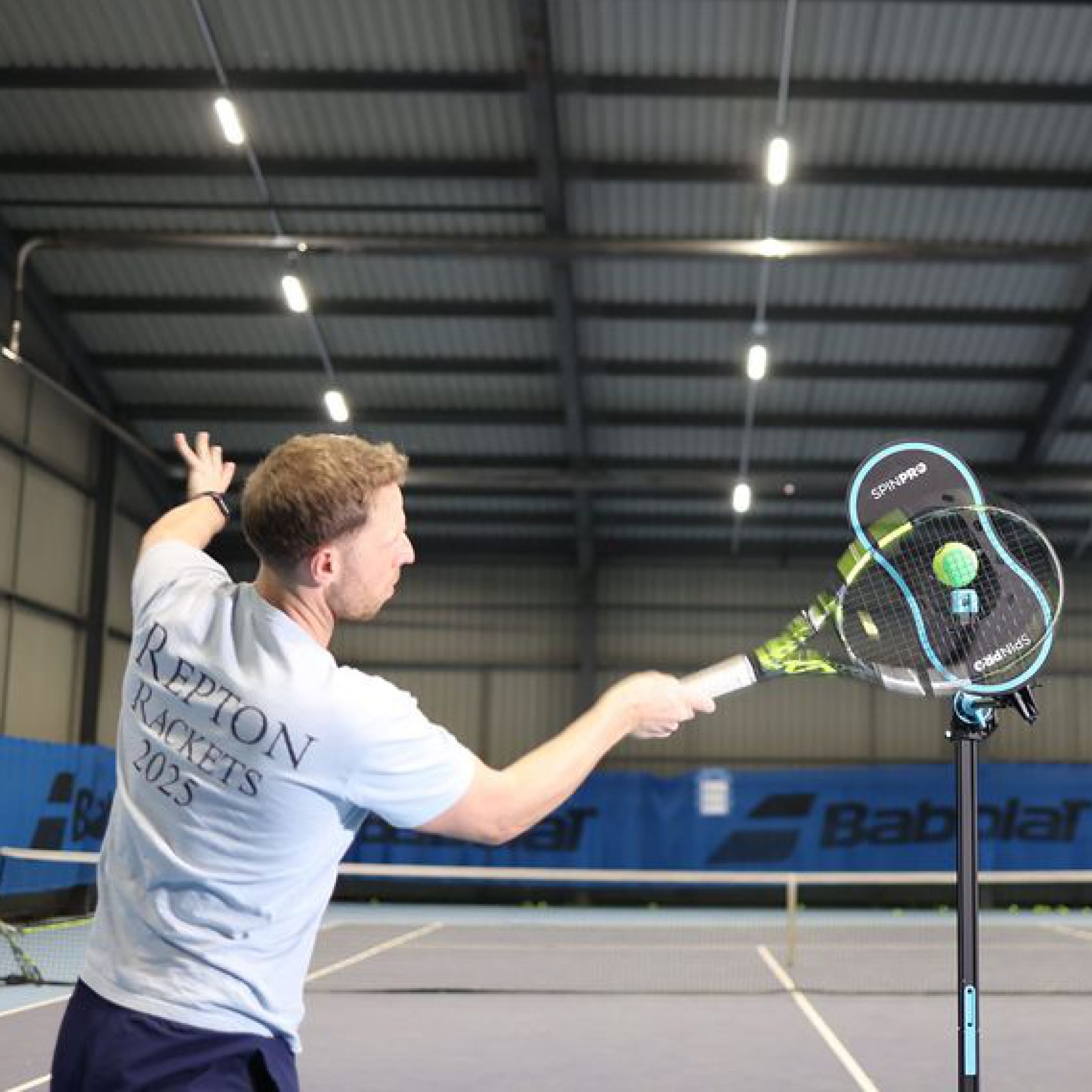 Player driving a high topspin forehand with the SpinPro on an indoor court, viewed from behind